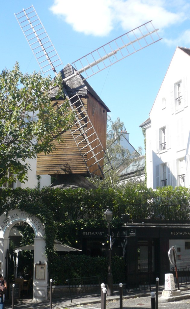 Eine Windmühle, mehrere Gebäude, Bäume, Metallstäbe und ein Zaun in einer ländlichen Landschaft.
