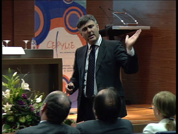 Ein Mann in einem schwarzen Blazer steht an einem Podium mit Mikrofonen, spricht in ein Handmikrofon, mit einem Blumentopf oder einem Blumenstrauß in der Nähe und einem Tisch mit einem Buch, einer Wasserflasche und anderen Gegenständen daneben, während drei Personen in Stühlen davor sitzen, mit einer braunen Wand und einer bunten Tafel mit Text im Hintergrund.