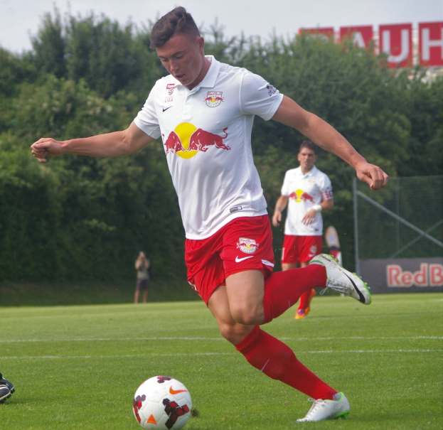 Professioneller Fußballspieler kickt einen Ball auf einem grünen Feld mit "RB Leipzig"-Schriftzug im Hintergrund unter einem klaren blauen Himmel.