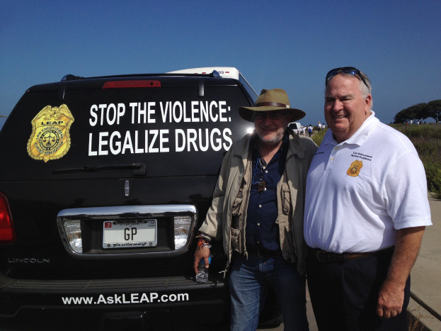 Zwei Männer stehen neben einem schwarzen Van mit 'Stop the Violence Legalize Drugs' darauf, einer hölt eine Flasche, mit Menschen, Bäumen und einem klaren blauen Himmel im Hintergrund.