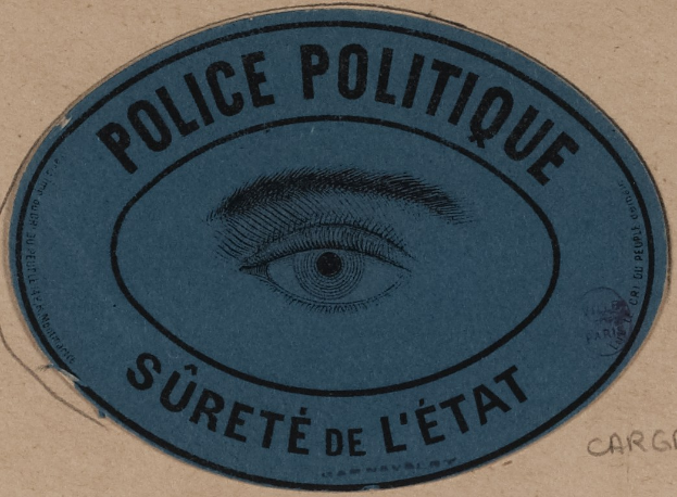 Braune Pappkiste mit blauem Aufkleber mit der Aufschrift "Polizei Politique Surete de L'Etat" und einem Auge in der Mitte.