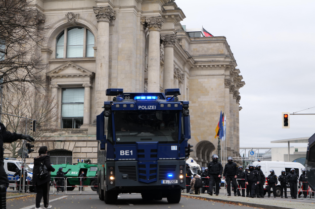 Eine Gruppe von Polizisten steht vor einem großen Gebäude mit Fenstern, Säulen und Bögen, mit Fahrzeugen auf der Straße und einer Person mit einer Kamera auf der linken Seite, Bäumen, Ampeln, Flaggen und einem klaren blauen Himmel im Hintergrund.