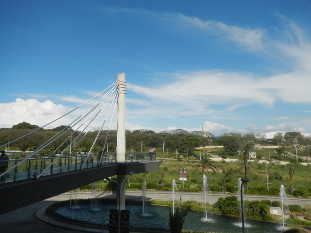 Eine Fußgängerbrücke mit Geländern und Säulen, umgeben von einem Brunnen in einem Park, mit Menschen, die darüber gehen, Laternenmasten und Schilder mit Text, vor einem Hintergrund aus Bäumen, Pflanzen, Gras, Hügeln und einem bewölkten Himmel.