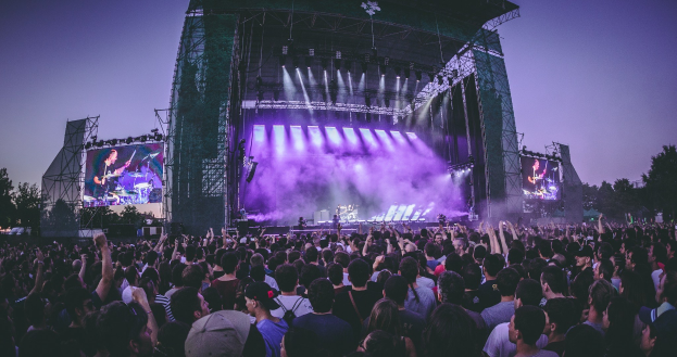 Eine große Menschenmenge auf einem Musikfestival schaut auf der hell erleuchteten Bühne mit Instrumenten und Bildschirmen, die vor einem Gebäude, Bäumen und einem blauen Himmel aufgebaut sind.