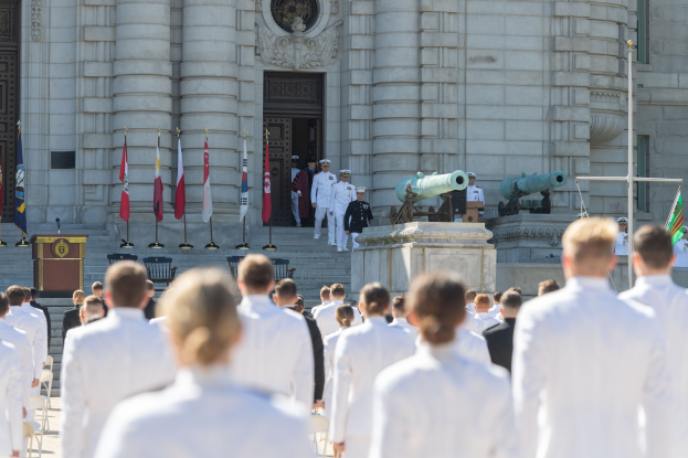 Gruppe von Menschen in weißen Marineuniformen auf einer Treppe vor einem Gebäude mit Säulen, Fahnen, einem Podium und Kanonen während einer Abschlussfeier.