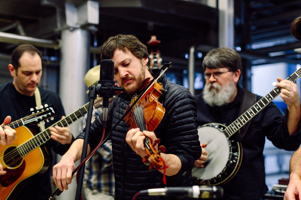 Eine Gruppe von Musikern in schwarzer Kleidung, die Instrumente spielen, mit einem Geiger am Mikrofon und einem Gebäude im Hintergrund.