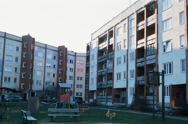 Ein Wohnhaus mit einem Spielplatz davor, der von Gras, Bänken, Schaukeln und anderen Gegenständen umgeben ist, mit Fahrzeugen, Bäumen und einem klaren blauen Himmel im Hintergrund.