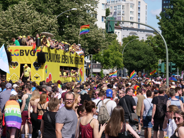 Eine große Menschenmenge marschiert auf einer Straße in Berlin, Deutschland, mit einem gelben Lastwagen im Hintergrund. Viele der Menschen tragen Mützen und Schutzbrillen und halten Fahnen. Es gibt Laternenpfähle und Ampeln mit Pfosten, sowie Bäume und Gebäude mit Fenstern. Der Himmel ist bewölkt und die Szene stammt wahrscheinlich von einem Christopher Street Day.