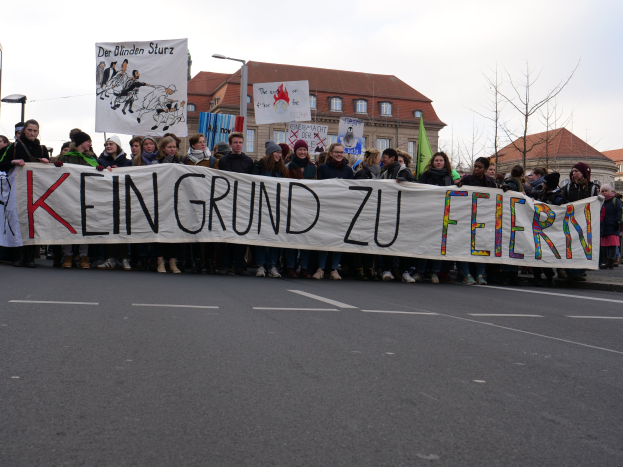 Eine Gruppe von Menschen mit einem Transparent mit der Aufschrift "Kein Grund zu Feiern", die gegen die deutschen Regierungskostenmaßnahmen protestiert, auf einer Straße mit Pfählen, Laternen, Bäumen, Gebäuden und einem klaren Himmel.