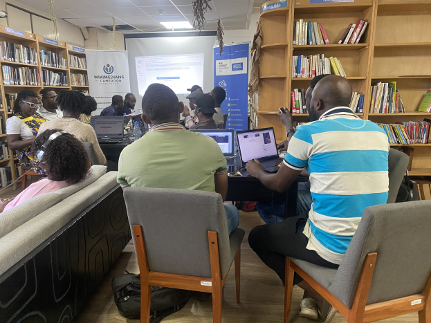 Gruppe von Menschen an einem Tisch mit Laptops umgeben von Bücherregalen in einer Bibliothekswerkstatt mit einem Banner, Bildschirm, Deckenleuchten und Taschen auf dem Boden.
