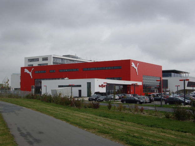 Fabrikgebäude von Puma in Deutschland mit Fahrzeugen auf der Straße, Straßenmasten, Laternen, Grünflächen und einem bewölkten Himmel.