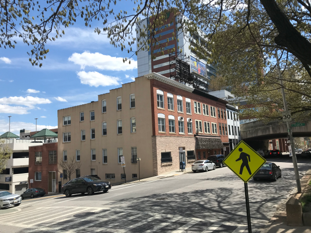 Eine Stra├če in einer Stadt mit parkenden Autos, Geb├Ąuden, Laternen, einem Schild f├╝r "Riverside Apartments in Nashville, TN", B├Ąumen, einer Br├╝cke und einem bew├Âlktem Himmel.