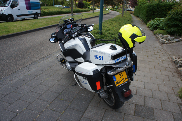 Ein Polizei-Motorrad am Straßenrand geparkt mit einem Helm darauf, mit Bäumen, Pfählen, Gebäuden, Pflanzen, Gras, Steinen und anderen Fahrzeugen im Hintergrund.