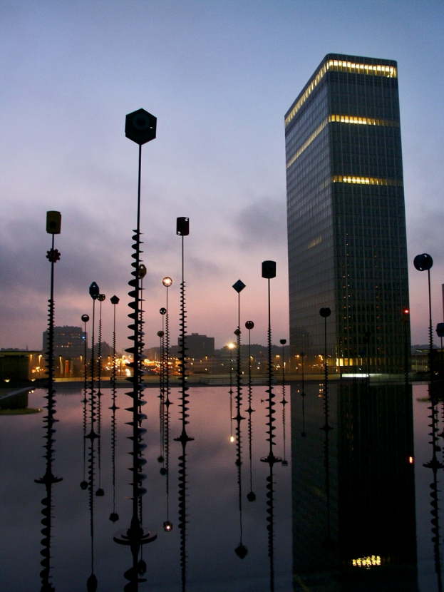 Mehrere Pfähle und Gebäude mit Lichtern unter einem bewölkten Himmel, mit ihren Spiegelungen