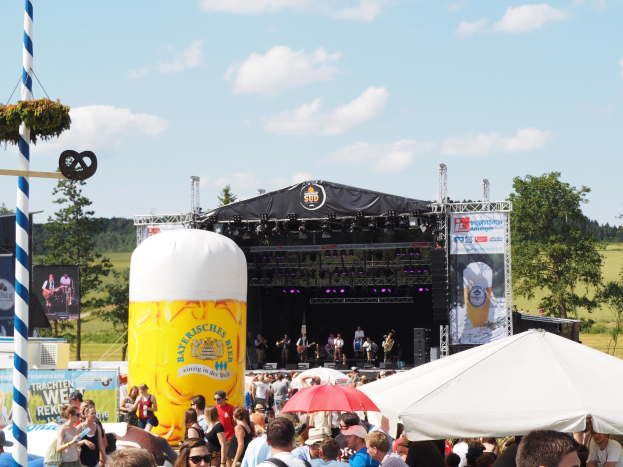 Eine große Menge beim Oktoberfest in München, Deutschland, vor einer Bühne mit Musikern, viele tragen Mützen und Schutzbrillen, einige halten Schirme, mit einem dekorierten Pfahl und Zelt im Vordergrund.