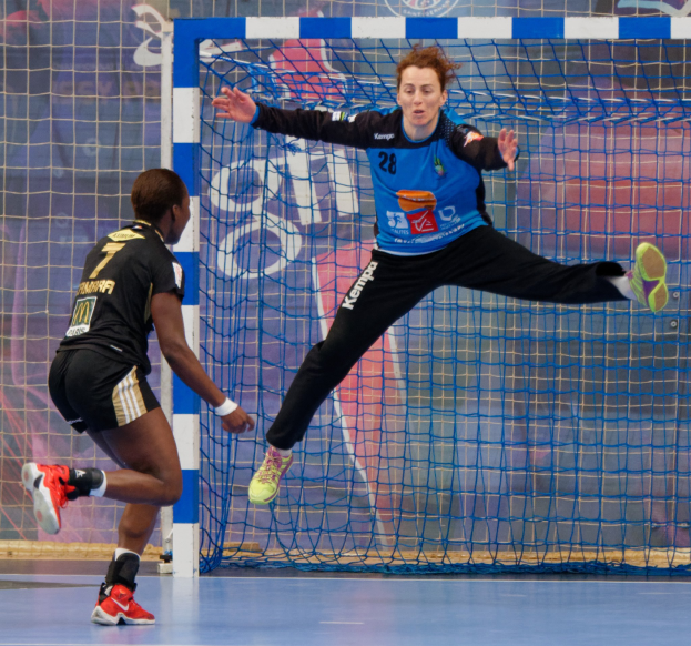 Zwei Frauen beim Handballspielen auf einem Platz mit einem Tor und einer Fahne im Hintergrund, beide tragen sportliche Schuhe.