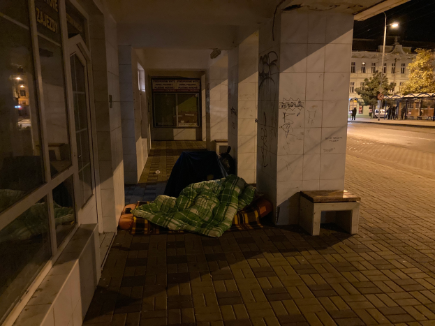 Eine Person schläft auf einer Decke auf dem Gehweg in der Nacht, mit einer Bank in der Nähe, während eine Gruppe von Menschen auf dem Gehweg steht, Fahrzeuge auf der Straße sind und Gebäude mit Fenstern im Hintergrund zu sehen sind unter einem bewölkten Himmel.