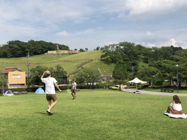 Eine Gruppe von Menschen, die Badminton in einem Park spielt, mit einem Mann, der einen Schläger hält und auf dem Gras in der Nähe von Zelten und Gebäuden sitzt.
