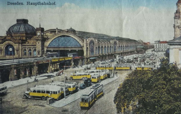 Eine alte Postkarte zeigt einen belebten Dresdner Bahnhof mit Fahrzeugen, Menschen, Bäumen, Gebäuden und einer bewölkten Himmel.