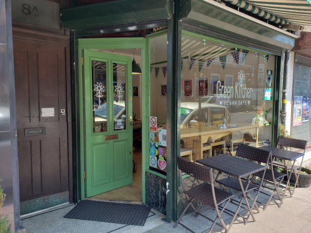 Ein Außenbereich mit Tischen und Stühlen bei Green Kitchen Vegan Cafe, umgeben von Pflanzen und einer Matte, mit einer Glaswand, die Plakate zeigt und einen Blick auf den Innenbereich mit einer hängenden Lampe gewährt.