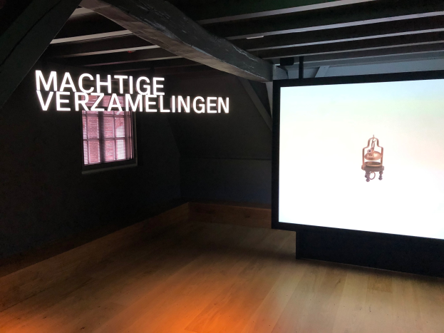 Installationsansicht der Ausstellung 'Machtige Verzamelingen' im Museum of Modern Art in Berlin, mit einem großen Bildschirm mit Wandtext und einem Fenster links.