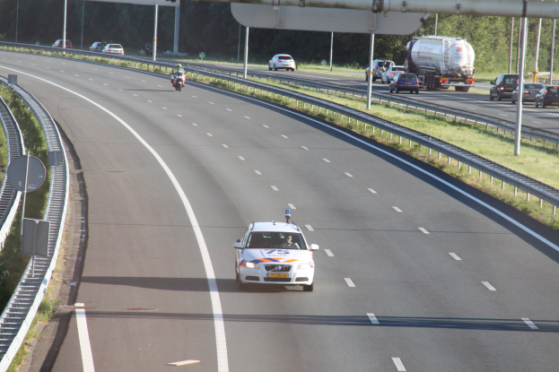 Ein Auto auf einer Autobahn mit einem stationären Polizeiwagen am Rande, andere Fahrzeuge und Bäume, Geländer, Pfosten und Planken im Hintergrund.