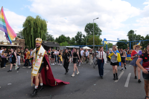 Eine Gruppe von Menschen marschiert bei der Gay Pride Parade 2018 mit einer Regenbogenflagge und Musikinstrumenten, im Hintergrund sind Laternenpfähle, Bäume, Schuppen und ein bewölkter Himmel zu sehen.