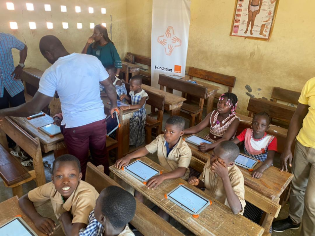 Eine Gruppe von Kindern sitzt an Tischen in einem Klassenzimmer und nutzt Tablets, umgeben von ein paar Menschen, sowie einem Banner und einem Plakat im Hintergrund.