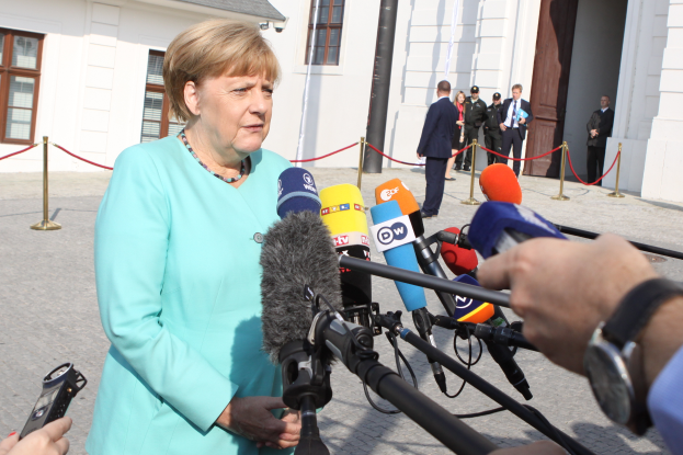 Bundeskanzlerin Angela Merkel spricht vor einem Gebäude mit Fenstern und einer Tür zu den Medien, umgeben von einer Gruppe von Menschen, mit einer Kamera und einem Handy auf der rechten Seite und Pfählen mit Seilen.