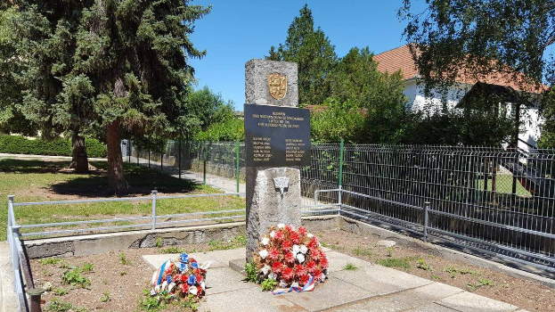 Ein Denkmal für die Opfer der Tschernobyl-Katastrophe, umgeben von einem Metallzaun, Kränzen, Gras, Pflanzen, Bäumen, mit einem Haus und einem bewölkten Himmel im Hintergrund.