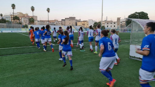 Eine Gruppe von Frauen, die Fußball auf einem Outdoor-Feld spielen, mit Gebäuden, Bäumen und einem Tor im Hintergrund.