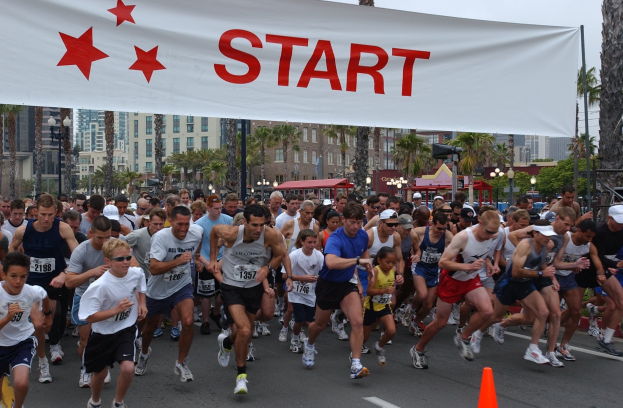 Eine Gruppe von Läufern bei einem Marathon läuft an einem Verkehrskegel vorbei, mit einem Banner und Gebäuden im Hintergrund bei klarem blauem Himmel.