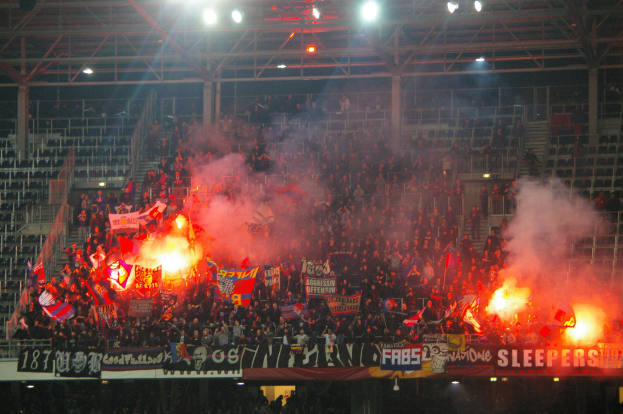 Eine gro├če Menge im Stadion h├Ąlt Fahnen und Banner, mit Rauchern, unter einer Decke mit Leuchten und Metallrahmen.