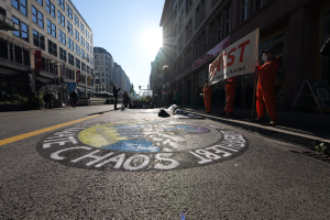Menschen mit Schildern und Plakaten stehen auf einer Straße, die mit Pflanzen und Gebäuden gesäumt ist, mit "Chaos" auf der Straße gemalt, in Anspielung auf die Occupy Wall Street-Proteste.