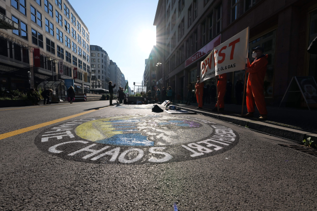 Menschen mit Schildern und Plakaten stehen auf einer Straße, die mit Pflanzen und Gebäuden gesäumt ist, mit "Chaos" auf der Straße gemalt, in Anspielung auf die Occupy Wall Street-Proteste.