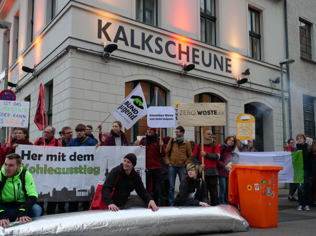 Eine Gruppe von Menschen mit Protestschildern und Plakaten vor einem Gebäude, mit zwei Personen im Vordergrund und einem Mülleimer rechts.