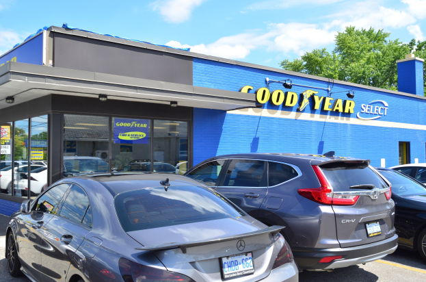 Außenansicht eines Goodyear Select-Autohauses mit mehreren parkenden Autos, einem Gebäude mit Text und Bäumen im Hintergrund sowie einem bewölkten Himmel.