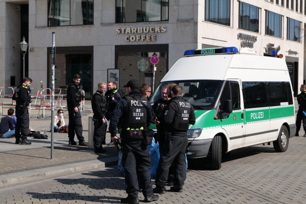 Eine Gruppe von Polizeibeamten steht vor einem Starbucks-Café, rechts davon ein Van und links ein paar Menschen; im Hintergrund ist ein Gebäude mit Fenstern, ein Schild, ein Laternenmast und ein Zaun zu sehen.