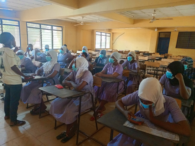 Ein Klassenraum in Nigeria mit Schülerinnen und Schülern, die Gesichtsmasken tragen und auf Bänken oder Stehplätzen sitzen, mit Büchern und Gegenständen auf Tischen, Fenstern, einer Tür und Deckenleuchten und -ventilatoren im Hintergrund.