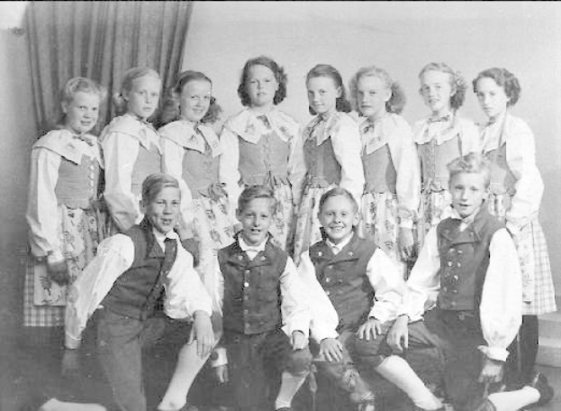 Schwarz-weiß-Foto einer Gruppe von Kindern, die vor einer Wand mit einem Vorhang posieren, mit der Beschriftung "1930er Kinder Schule" unten.