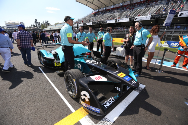 Eine Gruppe von Menschen feiert um ein Formel E-Rennwagen auf einer Rennstrecke, mit einem Stadion voller Zuschauer im Hintergrund.
