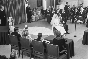 Schwarzes und weißes Foto einer Hochzeitszeremonie mit sitzenden und stehenden Menschen, einem Mann mit Mikrofon, einer Frau mit Blumenstrauß, einem Podium mit Mikrofon rechts und Blumenschmuck und Vorhang links.