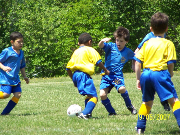 Kinder spielen Fussball auf einem grünen Feld mit Bäumen im Hintergrund und einem Datum in der rechten unteren Ecke.