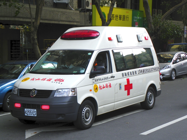 Ein Krankenwagen steht am Straßenrand geparkt, umgeben von anderen Fahrzeugen, mit Bäumen, Gebäuden, Texttafeln und Klimaanlagen im Hintergrund.