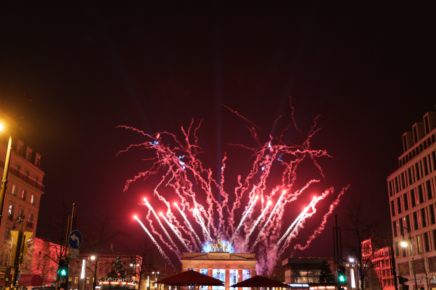 Eine belebte Straßenkreuzung während einer Silvesterfeier in Berlin, voller Menschen, Fahrzeuge und Gebäude, beleuchtet von Stadtlichtern und Feuerwerk am Himmel.