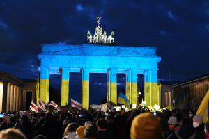 Eine Menschenmenge steht vor dem Brandenburger Tor in Berlin, hält Fahnen und Schilder, mit einer Banner auf der rechten Seite des Bildes.