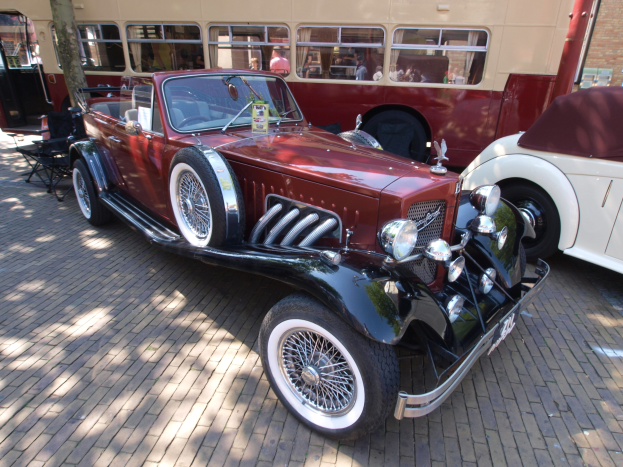 Ein Oldtimer neben einem Doppeldeckerbus auf einem Kopfsteinpflasterweg geparkt, mit Stühlen und einem Baum im Vordergrund, einem Gebäude mit Fenstern im Hintergrund und Menschen im Bus zu sehen.
