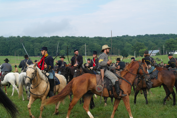 Eine Gruppe von Menschen zu Pferd mit Schwertern in der Vordergrund, Gras im Vordergrund und Bäume, Gebäude und Himmel im Hintergrund.