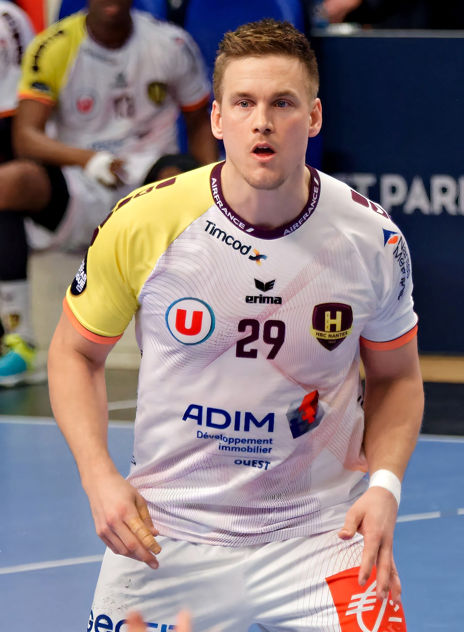 Ein Mann in weißem Hemd und gelben Shorts spielt Handball, hält den Ball und hat einen entschlossenen Gesichtsausdruck, während im Hintergrund Menschen auf dem Boden sitzen und eine Tafel mit der Aufschrift "Ligue 1 - Futsal - Olympique de Marseille" zu sehen ist.