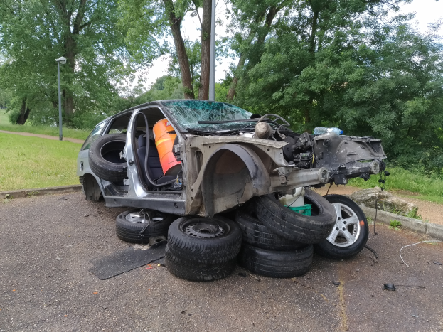 Ein Auto liegt umgekippt am Straßenrand, umgeben von Gras, Pflanzen, Bäumen, einer Straßenlaterne und einem bewölkten Himmel, mit herumliegenden Trömmern, die auf einen Unfall hindeuten.